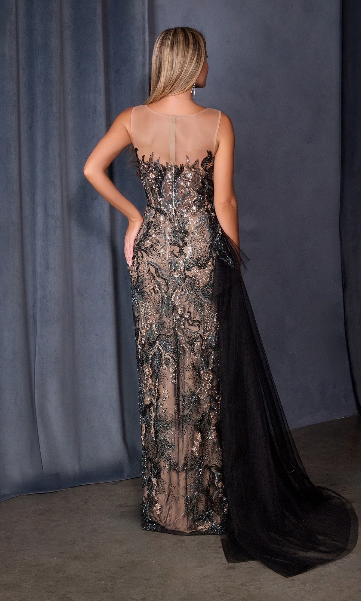 A woman faces away from the camera, wearing the Couture Long Black Prom Dress Andrea & Leo A1489 with a sheer-illusion neckline, sequin embroidery, and a flowing tulle train, set before dark draped curtains.